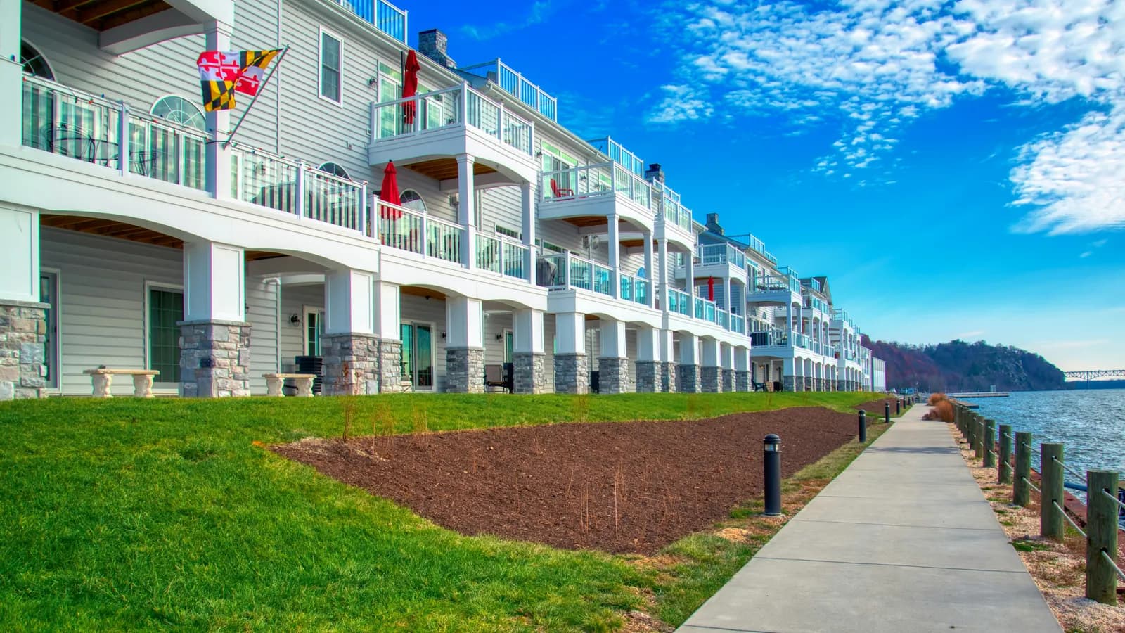 A row of waterfront townhouses with stacked balconies, stone-based columns and a lawn under a blue sky.